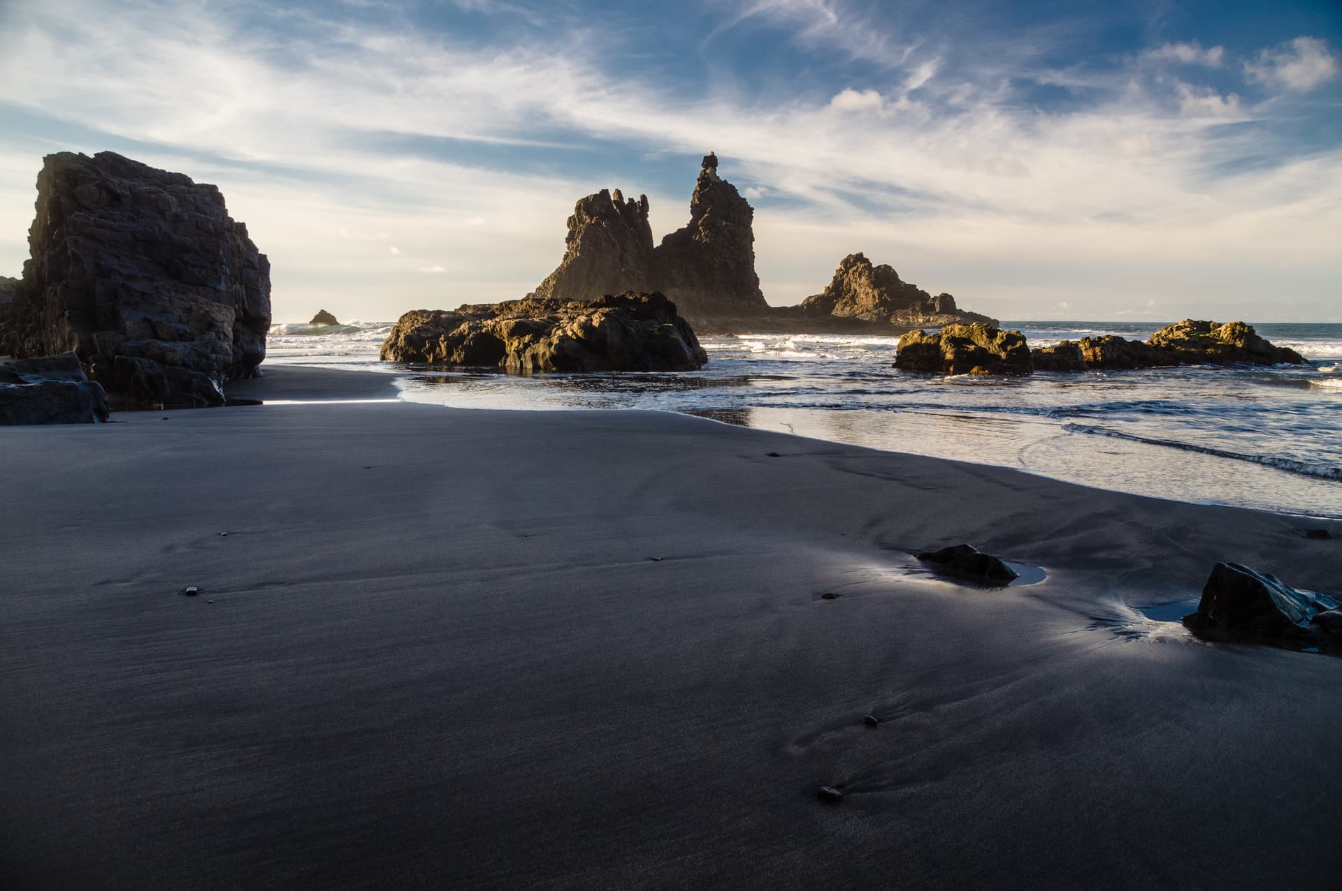 Rocky coast of Benijo beach with dark sand, sea stacks, and ocean waves under a cloudy sky.
